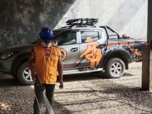An exterminator from Technologist Pest Control stands indoors on a gravel floor, wearing a blue hard hat, yellow safety vest, respirator mask, and holding a spraying device. Behind him is a black service pickup truck featuring bold graphics of a person spraying, a scorpion logo, and the contact number “0927-365-2847”—signaling an active pest control operation.