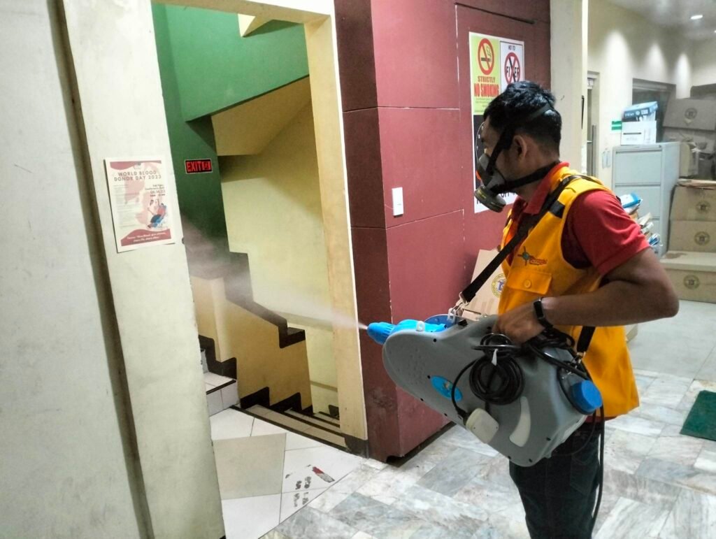 Alt Text for Techno Pest Control Disinfection Image A sanitation specialist from Techno Pest Control, wearing a yellow safety vest and red shirt, uses a fogging machine to disinfect a stairwell in a public facility. Green and beige walls frame the tiled area as posters on health and safety, including one for "World Blood Donor Day 2023," emphasize the environment’s importance. An "EXIT" sign glows above the stairs.