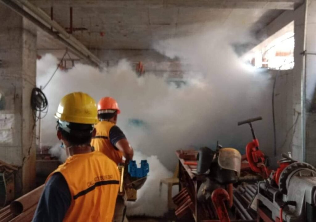 Two pest control technicians OF TECHNO pEST cONTROL wearing safety helmets and orange vests are conducting a fogging operation inside an unfinished building or construction site. Thick white fog, likely insecticide, fills the air to eliminate flying insects such as mosquitoes. The area is filled with construction materials and tools, highlighting the need for pest control even during building phases.