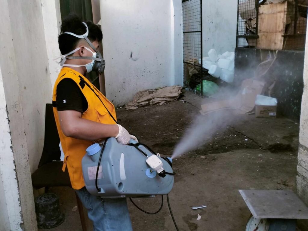 Technologist Pest Control technician fogging a narrow alley with misting equipment, wearing safety gear and mask to disinfect and control mosquito or pest populations in an urban outdoor setting.